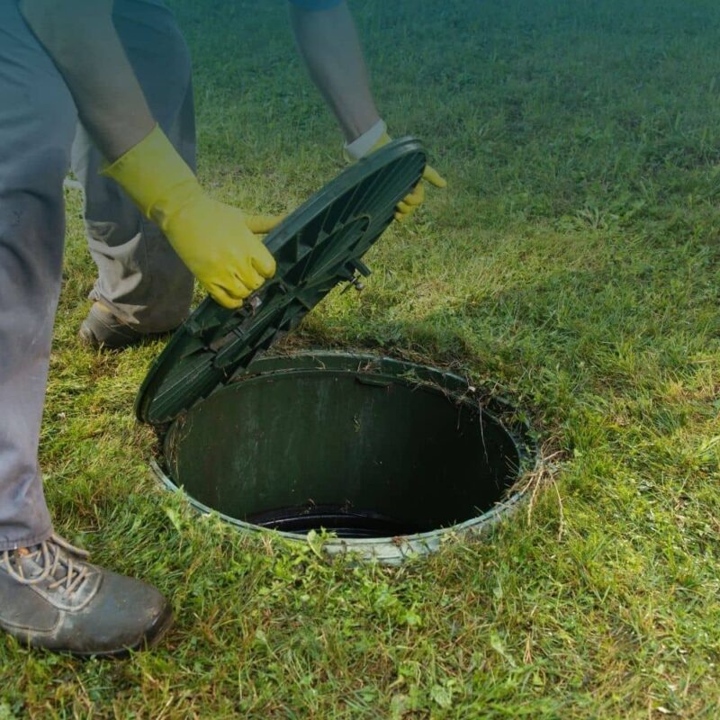 how to locate a septic tank: a person's gloved hands lifting a round septic tank access lid out of the ground