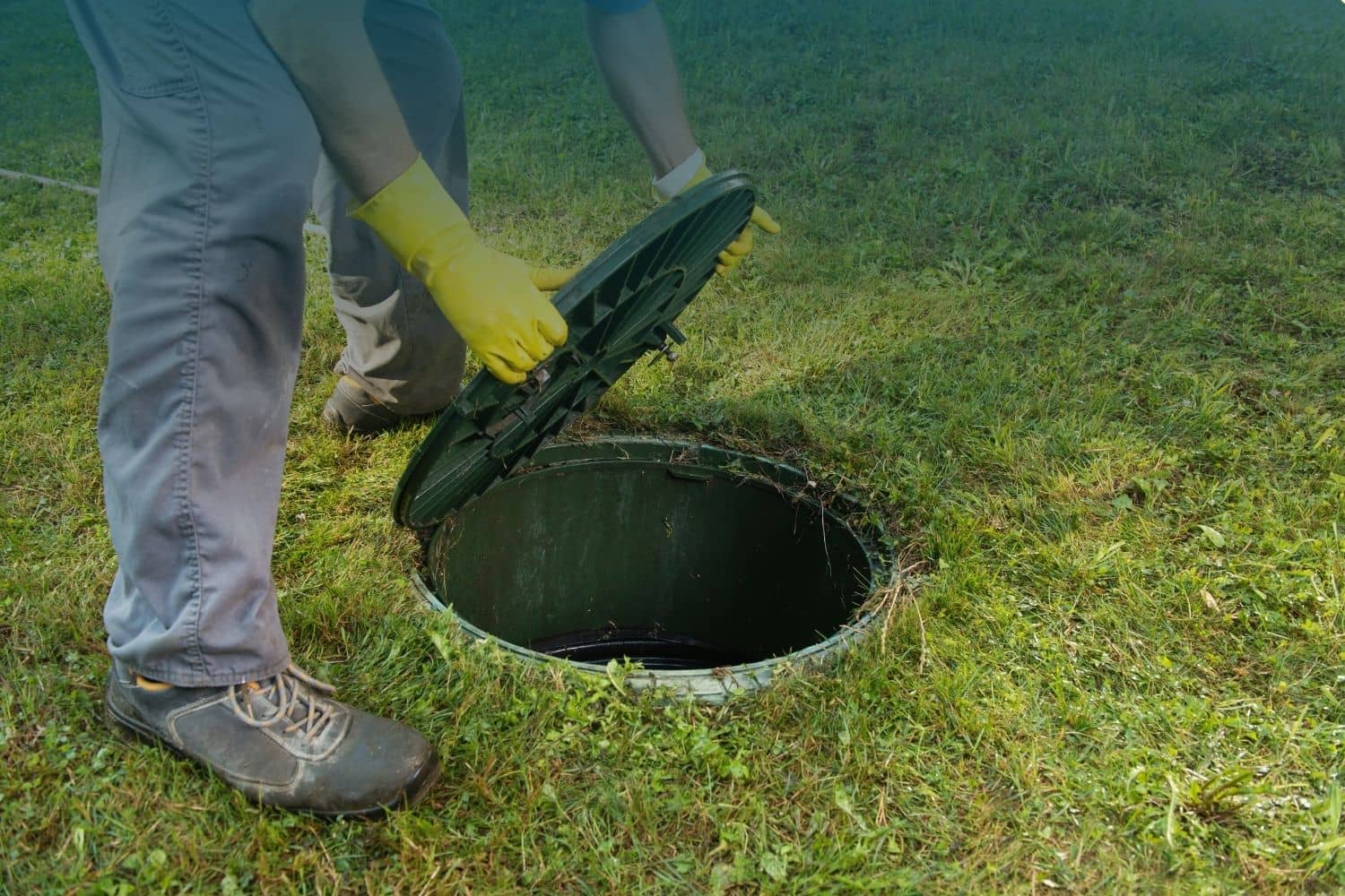 how to locate a septic tank: a person's gloved hands lifting a round septic tank access lid out of the ground