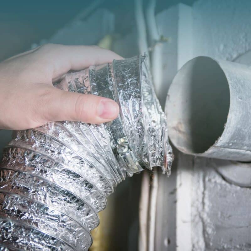 A hand connects a shiny, flexible aluminum duct to a circular wall dryer vent. The duct appears newly installed, conveying a sense of maintenance or repair.