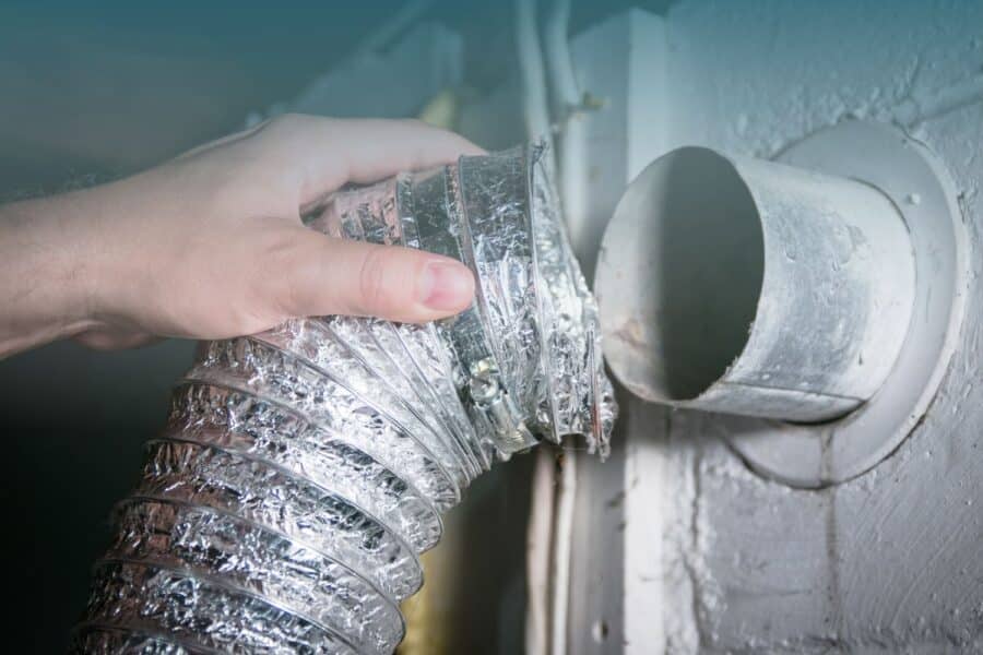 A hand connects a shiny, flexible aluminum duct to a circular wall dryer vent. The duct appears newly installed, conveying a sense of maintenance or repair.