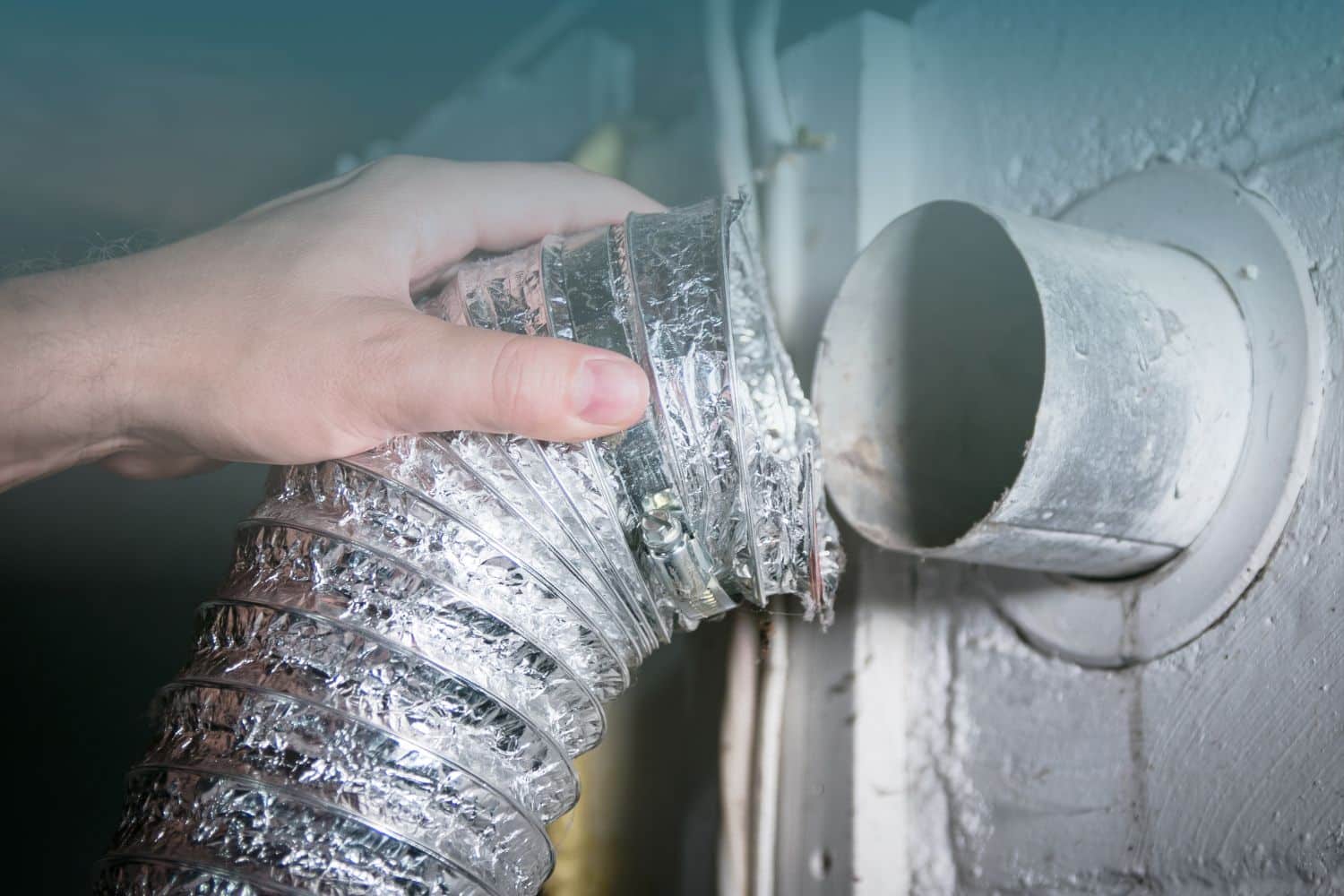 A hand connects a shiny, flexible aluminum duct to a circular wall dryer vent. The duct appears newly installed, conveying a sense of maintenance or repair.